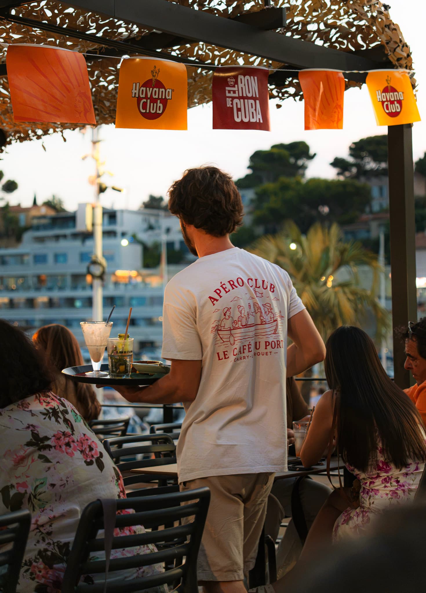 Un serveur en t-shirt Apéro Club apporte les boissons sur la terrasse